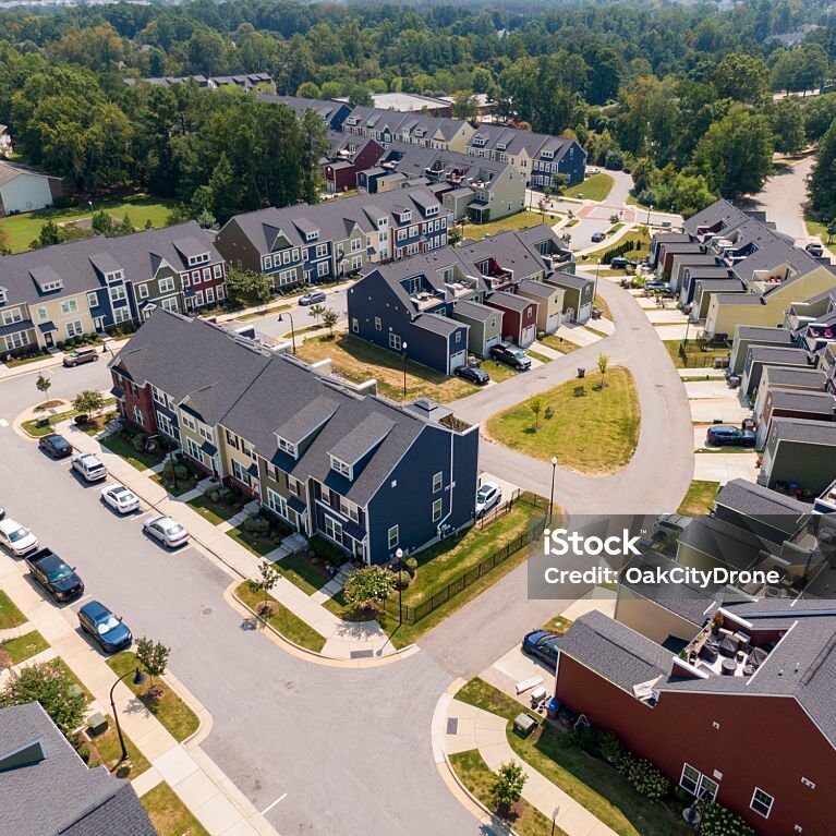 This professional aerial shot provides a comprehensive view of a contemporary multi-family residential development. The architecture is defined by staggered townhome units with a mix of colorful fiber cement siding and consistent dark gray architectural shingle roofing. The community layout includes clean asphalt roads, white-painted crosswalks, and small green common areas with young trees. Several parked vehicles are visible along the curbs and in private driveways, illustrating a quiet suburban lifestyle. This imagery is ideal for real estate marketing, urban planning, and housing development themes.